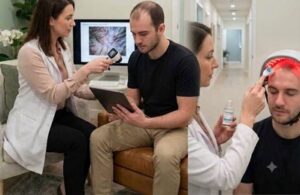 A female specialist examines a patient's scalp using a trichoscope while reviewing digital diagnostics during a non-surgical consultation.