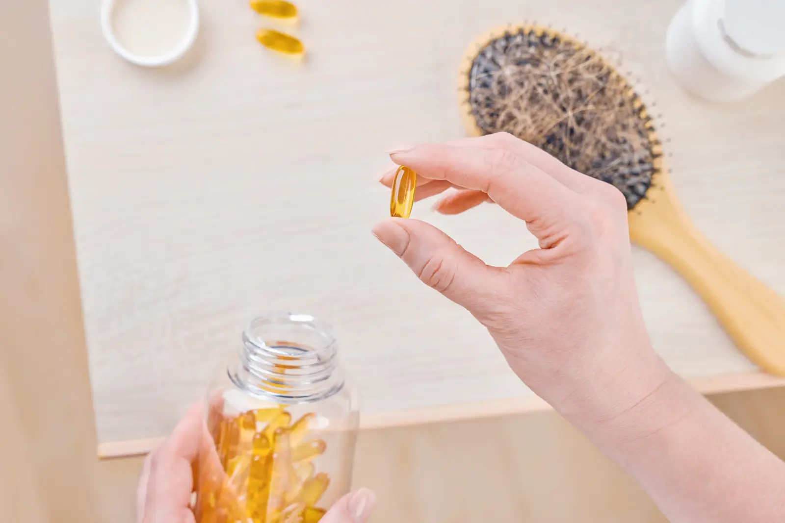 Hand holding a golden supplement capsule above an open bottle of soft gels, with a hairbrush full of shed hair in the background